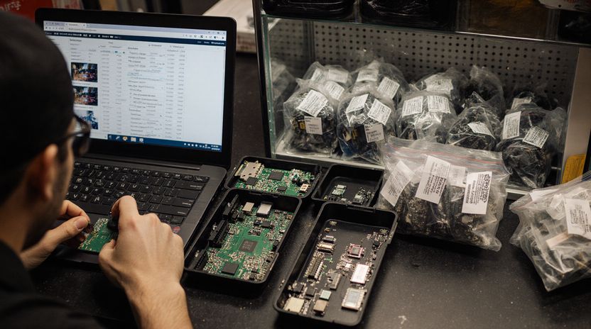 A technician at the bench with a circuit board in hand, more boards in trays beside the laptop