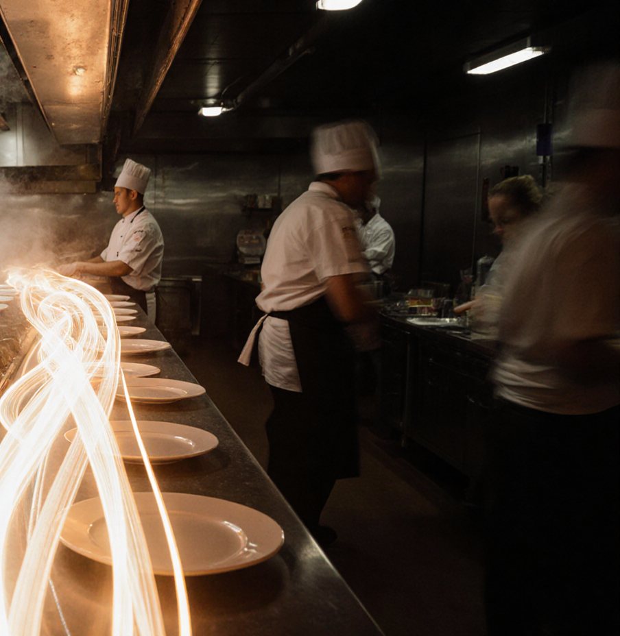 Cooks in white jackets and toques work a dim kitchen line, a row of white plates along the pass traced by ribbons of light in a long exposure