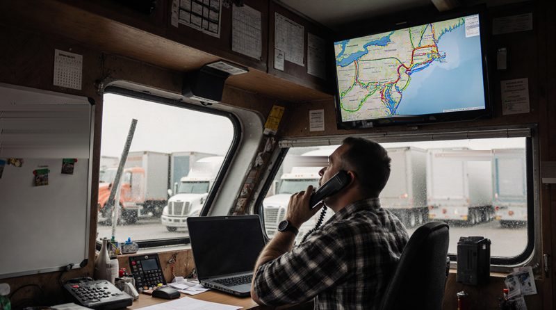 A dispatcher in a flannel shirt on the phone at his desk in a yard office, a laptop open in front of him and a wall screen above showing a road map of the Northeast with route lines; parked trucks and trailers outside the rain-streaked window